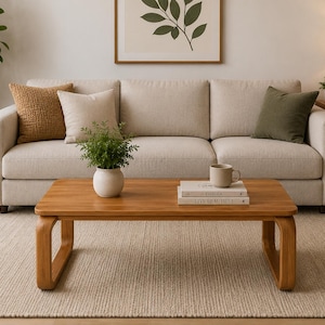 May include: A rectangular wooden coffee table with curved legs, featuring a light brown finish. On the table sits a small potted green plant, a beige ceramic mug, and two stacked books titled Kinfolk and Live Beautiful.