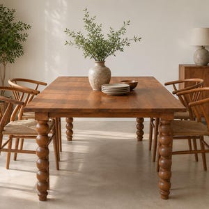 May include: A rectangular wooden dining table with four thick, stacked-sphere legs, accompanied by four wooden chairs featuring curved backrests and woven seats. A ceramic vase with green foliage and a stack of plates sit on the tabletop.
