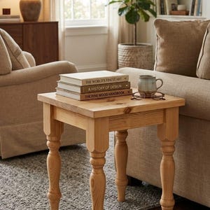 Puede incluir: Una mesa auxiliar cuadrada de madera clara con cuatro patas torneadas. Sobre la superficie de la mesa hay tres libros apilados titulados The World's Finest Art, A History of Design y The Pine Wood Handbook, junto a una taza y unas gafas.
