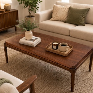 Puede incluir: Una mesa de centro rectangular de madera marrón oscuro con bordes redondeados y cuatro patas cónicas. Sobre la mesa hay un jarrón blanco acanalado con vegetación, dos libros apilados y una bandeja tejida con una vela y una taza de cerámica.