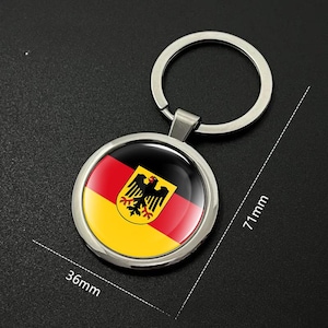Op de afbeelding: Een ronde zilveren sleutelhanger met een hanger van 36 mm diameter met de Duitse vlag. Het vlagontwerp omvat zwarte, rode en gele strepen met een zwart adelaarsymbool. De sleutelhanger is 71 mm lang.