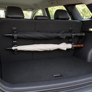 May include: A black and a beige umbrella held horizontally by two black plastic mounting clips attached to the back of a vehicle seat. The umbrellas are positioned one above the other against the dark grey fabric of the rear car seat interior.