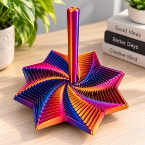 May include: A kinetic sculpture featuring a central vertical rod and a multi-layered, star-shaped base with a gradient finish transitioning from deep blue to vibrant purple, orange, and yellow, creating a complex, repeating geometric pattern.
