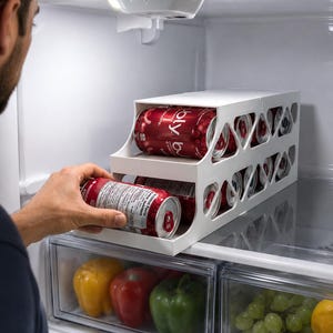 May include: A white plastic two-tier refrigerator beverage organiser rack with triangular side cutouts, holding multiple red aluminium soda cans. The organiser is designed to dispense cans from the bottom shelf while storing them on the top.