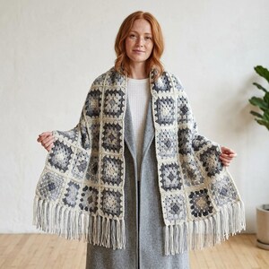 May include: A rectangular crochet shawl featuring a repeating granny square pattern in shades of charcoal gray, light gray, and cream. The shawl is finished with long, cream-colored fringe along the bottom edge, creating a textured appearance.
