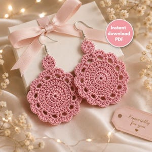 Puede incluir: Un par de pendientes de ganchillo en color rosa empolvado con un diseño circular festoneado y ganchos de metal plateado. Los pendientes se presentan sobre una caja de regalo blanca con un lazo rosa y una etiqueta que dice Especially for you.