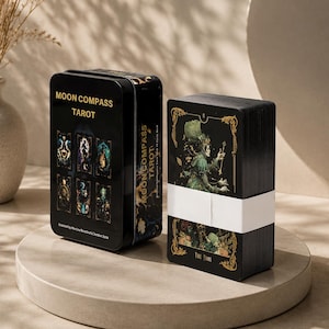 Może przedstawiać: Czarna prostokątna metalowa puszka z napisem Moon Compass Tarot i sześcioma ilustracjami kart z przodu, obok talii kart tarota owiniętej białym papierowym paskiem. Na wierzchniej karcie widać postać w zielonym kapeluszu oraz napis The Fool.