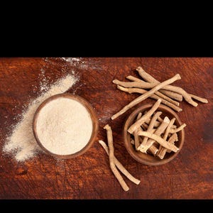 May include: Two small wooden bowls containing ashwagandha. One bowl is filled with a fine, light beige powder, with some spilled on the surface. The other bowl holds several dried, woody, light brown root segments arranged in a cluster.