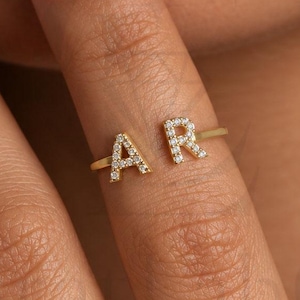 Puede incluir: Un anillo abierto de tono dorado con las letras mayúsculas A y R, incrustadas con pequeñas piedras transparentes. Las letras están situadas en los extremos de la banda metálica fina y pulida, creando un espacio entre ambos caracteres.