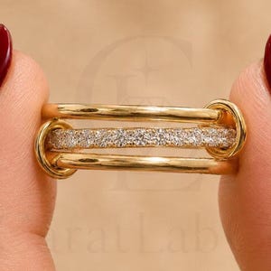 Könnte beinhalten: Ein Goldring bestehend aus drei parallelen Bändern, die an den Seiten durch zwei kreisförmige Schlaufen verbunden sind. Das mittlere Band ist mit einer Reihe kleiner, klarer, funkelnder Steine besetzt, während das obere und untere Band glatt sind.