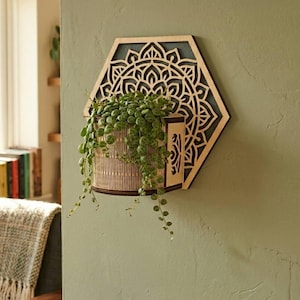 Puede incluir: Un macetero de pared hexagonal de madera con un diseño de mandala cortado con láser. El macetero contiene una planta verde colgante. El macetero está montado en una pared verde, creando un acento decorativo y natural.