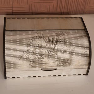 May include: Wooden bread box with a roll-top lid. The box features a laser-etched design of a baker with wheat and bread. The box is made of light-colored wood with dark brown trim. The design is detailed and adds a rustic touch.