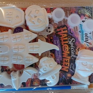 Peut inclure: Un kit créatif d'Halloween dans un sachet plastique transparent contenant des figurines en plâtre blanc représentant une maison hantée, une pierre tombale avec RIP, une citrouille, une chauve-souris, une momie et un chapeau de sorcière.