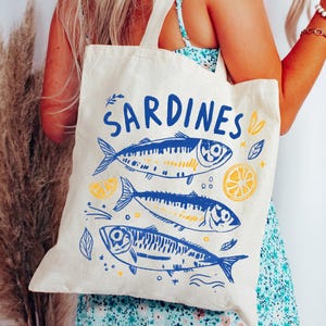 Könnte beinhalten: Eine cremefarbene Stofftasche aus Segeltuch mit einem blauen Grafikdruck von drei vertikal gestapelten Sardinen. Das Wort SARDINES ist in Blau über den Fischen aufgedruckt, ergänzt durch kleine gelbe Zitronenscheiben und blaue Blattmotive auf dem Stoff.