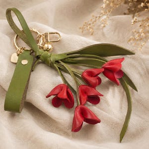 Puede incluir: Un llavero de cuero hecho a mano con un ramo de flores de tulipanes rojos. El llavero tiene una correa de cuero verde y herrajes dorados. Los p&eacute;talos de los tulipanes son de un rojo vibrante y las hojas son verdes.