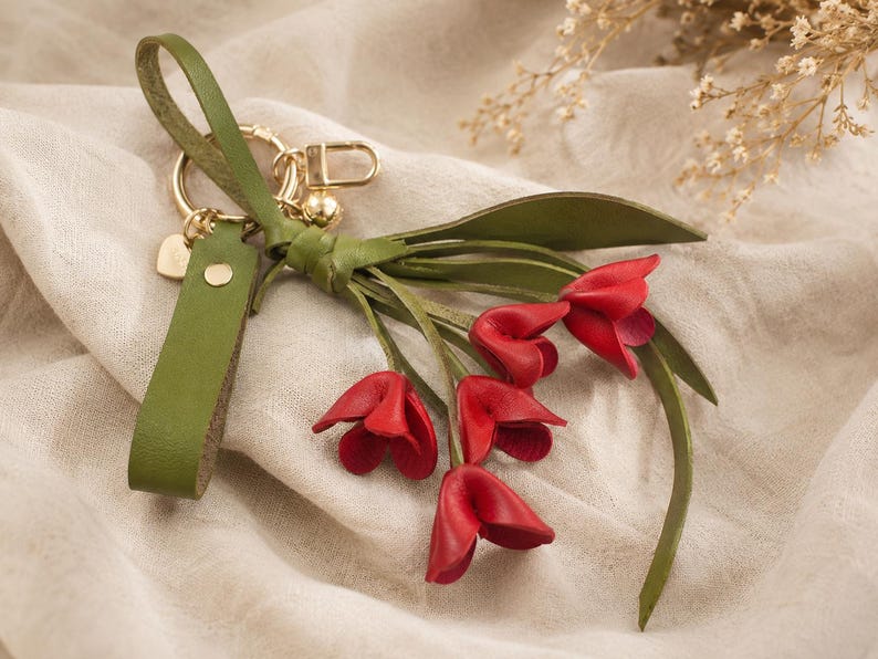 Puede incluir: Un llavero de cuero hecho a mano con un ramo de flores de tulipanes rojos. El llavero tiene una correa de cuero verde y herrajes dorados. Los p&eacute;talos de los tulipanes son de un rojo vibrante y las hojas son verdes.