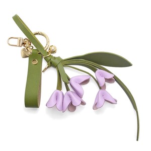 Puede incluir: Un llavero de cuero verde con un cierre dorado y un peque&ntilde;o dije en forma de coraz&oacute;n. El llavero presenta un grupo de capullos de flores de cuero de color lavanda con hojas verdes, creando un dise&ntilde;o floral.