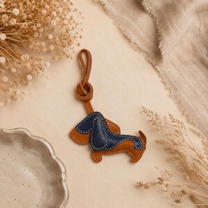 May include: A navy blue and tan leather dachshund keyring with white stitching. The keyring features a brown leather loop. The dog-shaped charm is a stylish accessory for keys or bags.