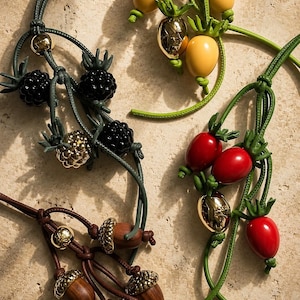 Peut inclure: Gros plan de plusieurs colliers avec des motifs de fruits et de glands. Les colliers présentent des fruits jaunes, rouges et noirs, ainsi que des glands bruns. Les fruits et les glands ont des accents dorés et sont enfilés sur des cordons verts et bruns.