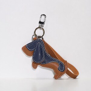 May include: A leather dachshund keyring in brown and navy blue with white stitching. The keyring has a silver-coloured clasp and a brown leather strap. The dog-shaped charm is approximately 7.6 cm long.