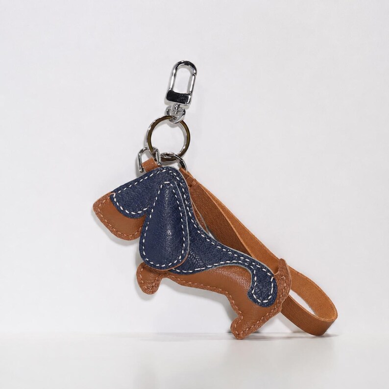 May include: A leather dachshund keyring in brown and navy blue with white stitching. The keyring has a silver-coloured clasp and a brown leather strap. The dog-shaped charm is approximately 7.6 cm long.