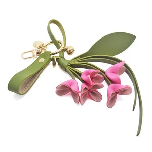 Puede incluir: Un llavero de cuero verde con un cierre y detalles dorados. El llavero presenta un grupo de flores de cuero rosa con tallos y hojas verdes. Tambi&eacute;n se adjuntan una peque&ntilde;a campana y un dije dorados.