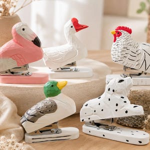 May include: A collection of five animal-shaped staplers featuring a pink flamingo, a white goose with a red beak, a white chicken with a red comb, a mallard duck with a green head, and a white dog with black spots, all with carved wood textures.
