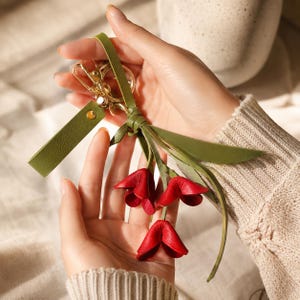 Puede incluir: Un llavero de cuero hecho a mano con un racimo de tres flores rojas en forma de tulip&aacute;n. El llavero tiene una correa de cuero verde y herrajes dorados. Las flores son de un rojo intenso y las hojas son verdes.
