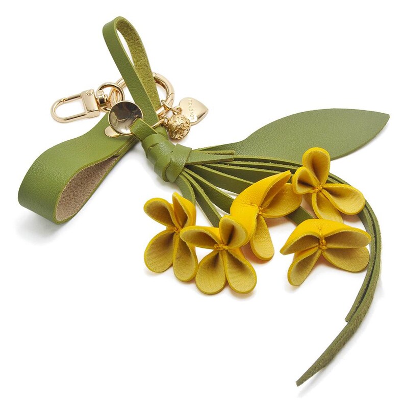 Puede incluir: Un llavero de cuero verde con cierre y herrajes dorados. El llavero presenta un grupo de flores amarillas y una borla de cuero verde. Un peque&ntilde;o dije de coraz&oacute;n dorado est&aacute; adjunto.