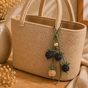 May include: A beige woven tote bag with a matching handle. The bag is decorated with a green string of beaded black and gold berry charms. The bag is sitting on a wooden surface.