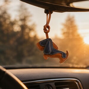 May include: A leather dachshund car charm in brown and navy blue, suspended from a car's rearview mirror. The charm is stitched with white thread and has a leather loop. The background is blurred, with a warm, golden light.