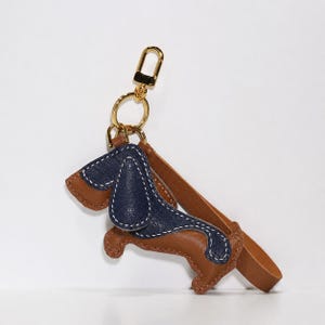 May include: A dachshund-shaped leather keychain in brown and navy blue, with white stitching. It features a gold-coloured clasp and a brown leather strap. The charm is dog-shaped.