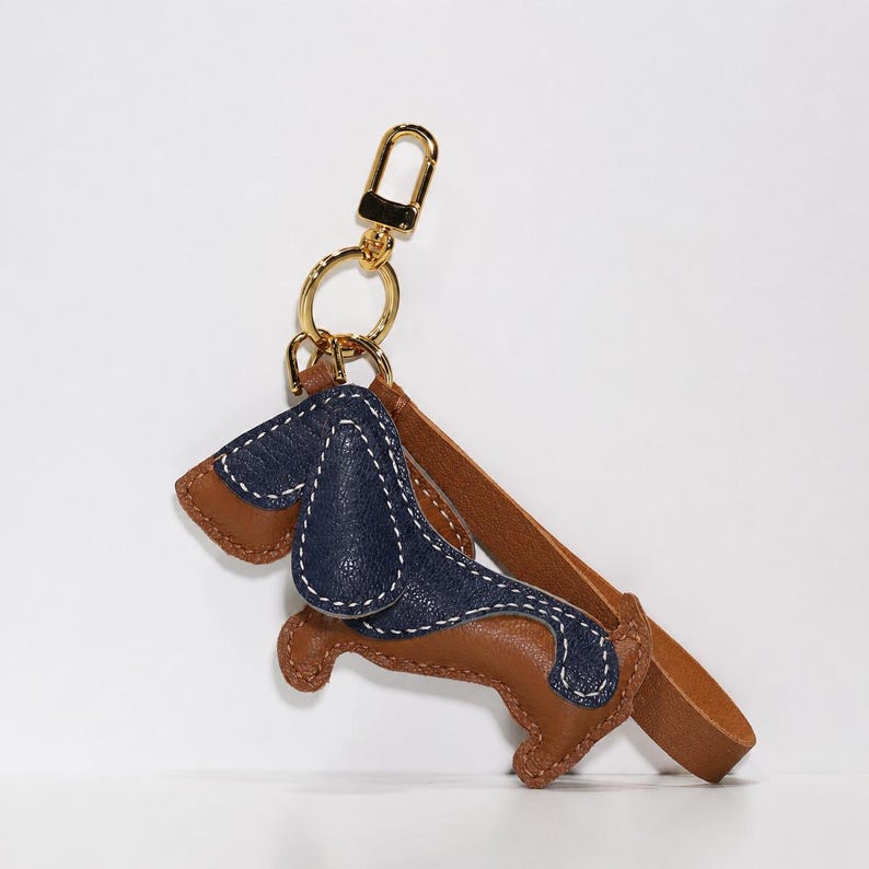 May include: A dachshund-shaped leather keychain in brown and navy blue, with white stitching. It features a gold-coloured clasp and a brown leather strap. The charm is dog-shaped.