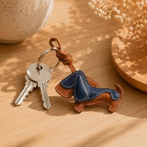 May include: A brown and navy blue leather dachshund keyring with white stitching. The keyring is attached to a silver ring with two keys. The dog-shaped charm has a leather loop for attachment.