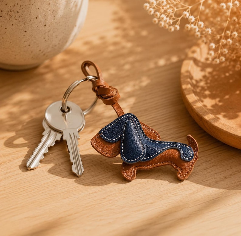May include: A brown and navy blue leather dachshund keyring with white stitching. The keyring is attached to a silver ring with two keys. The dog-shaped charm has a leather loop for attachment.