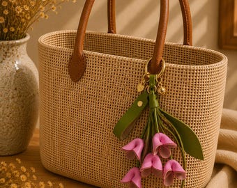 Puede incluir: Un bolso tote tejido con asas y detalles de cuero marrón. El bolso presenta un adorno floral verde y rosa decorativo. El bolso está sobre una superficie de madera.