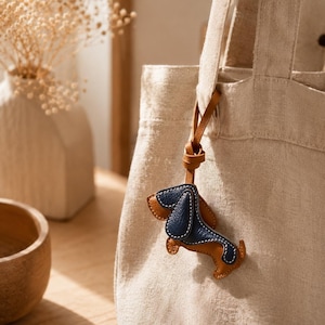 May include: A beige canvas tote bag with a brown leather strap, featuring a small leather dachshund dog charm. The charm is brown and navy blue with white stitching. A vase with dried flowers is in the background.