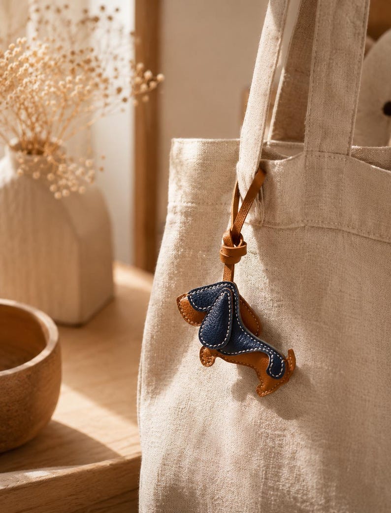 May include: A beige canvas tote bag with a brown leather strap, featuring a small leather dachshund dog charm. The charm is brown and navy blue with white stitching. A vase with dried flowers is in the background.