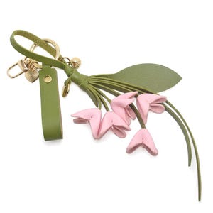Puede incluir: Un llavero de cuero verde con un cierre dorado y un dise&ntilde;o de flores rosas. El llavero presenta una correa verde, un dije en forma de coraz&oacute;n y un grupo de flores rosas con forma de tulip&aacute;n con tallos y hojas verdes.