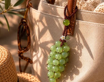Puede incluir: Un amuleto de racimo de uvas verdes con cuentas cuelga de un asa de cuero marrón de un bolso tote beige. El amuleto está sujeto con un cordón marrón y presenta una pequeña flor blanca y una cuenta verde. También se ven gafas de sol y un sombrero de paja.
