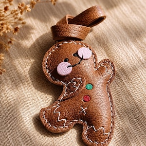 May include: A brown leather gingerbread man keychain with white stitching. The gingerbread man has pink circular eyes, a black smile, and a green and red button. A brown leather loop is attached to the top.
