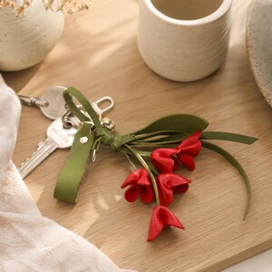 Puede incluir: Un llavero con una correa de cuero verde y un racimo de flores rojas con forma de tulip&aacute;n. El llavero est&aacute; junto a un juego de llaves, una taza de cer&aacute;mica blanca y un jarr&oacute;n decorativo sobre una superficie de madera.