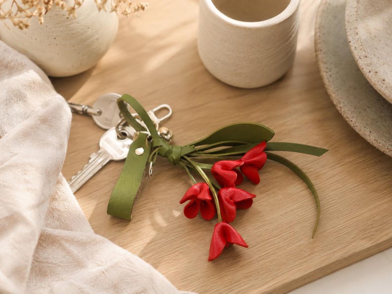 Puede incluir: Un llavero con una correa de cuero verde y un racimo de flores rojas con forma de tulip&aacute;n. El llavero est&aacute; junto a un juego de llaves, una taza de cer&aacute;mica blanca y un jarr&oacute;n decorativo sobre una superficie de madera.