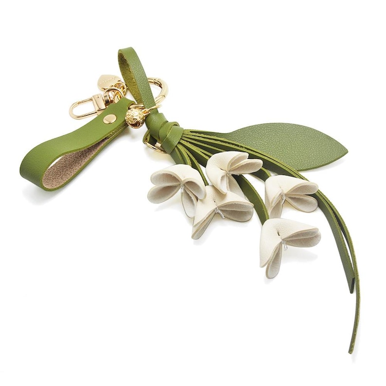 Puede incluir: Un llavero de cuero verde con un cierre dorado y un dise&ntilde;o floral decorativo. El llavero presenta una correa verde, una borla y varios acentos en forma de flor de color blanquecino. El dise&ntilde;o general es elegante e inspirado en la naturaleza.