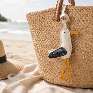 May include: A seagull-shaped leather bag charm featuring a white body, a dark blue wing with yellow stitching, a yellow beak, and dangling yellow feet. The charm is attached to a woven straw tote bag with brown leather handles.
