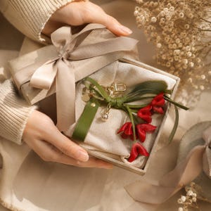 Puede incluir: Una caja de regalo envuelta en papel beige y atada con una cinta de sat&eacute;n. En el interior, un llavero de cuero con detalles de tulipanes rojos y una correa de cuero verde se presenta sobre una tela color crema.