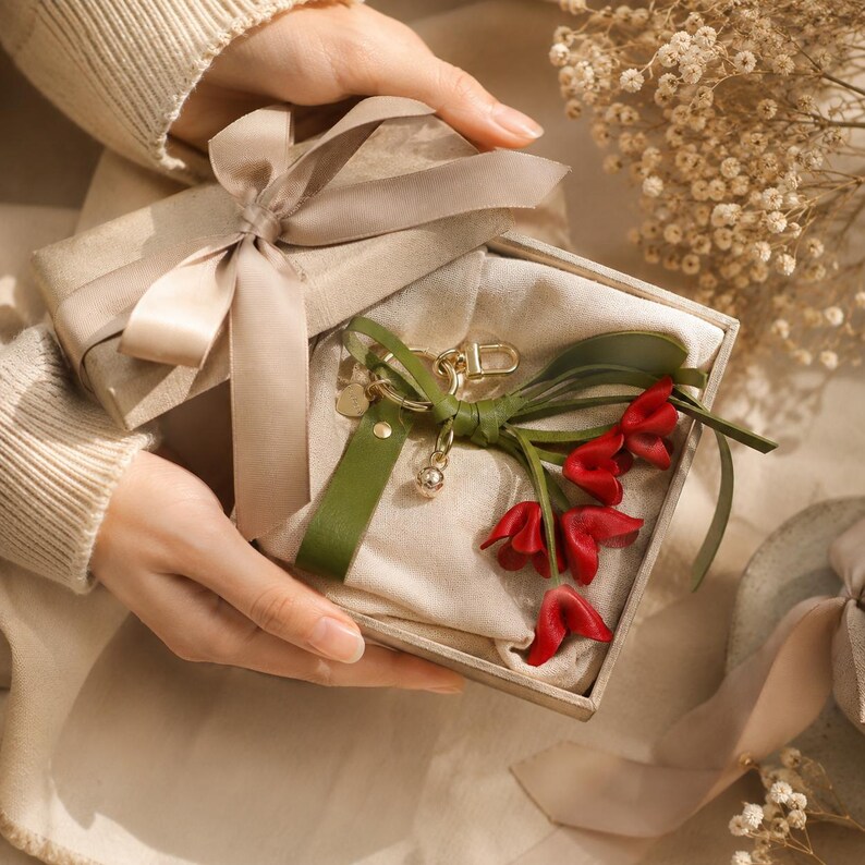 Puede incluir: Una caja de regalo envuelta en papel beige y atada con una cinta de sat&eacute;n. En el interior, un llavero de cuero con detalles de tulipanes rojos y una correa de cuero verde se presenta sobre una tela color crema.