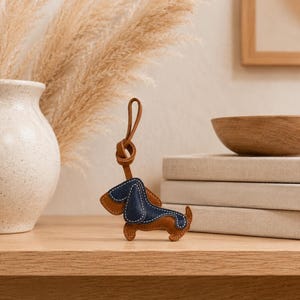 May include: A brown and navy blue leather dachshund dog keyring with a brown leather loop. The dog has a stitched navy blue back and ears, with brown leather on the legs and body. The keyring is on a wooden surface.