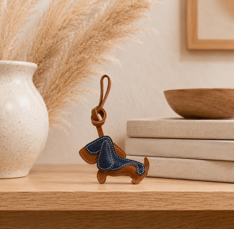 May include: A brown and navy blue leather dachshund dog keyring with a brown leather loop. The dog has a stitched navy blue back and ears, with brown leather on the legs and body. The keyring is on a wooden surface.
