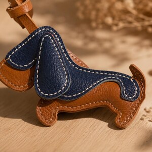 May include: A brown and navy blue leather dog-shaped keyring. The keyring has white stitching and a brown leather strap. The dog's ears and back are navy blue, while the body is brown. The keyring is on a wooden surface.